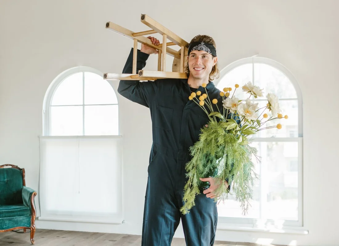 Mann trägt einen Holzstuhl und eine Blumenvase durch einen hellen Wohnraum.